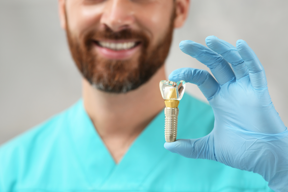 Dentist holding a dental implant with crown in blue gloves, smiling at camera.